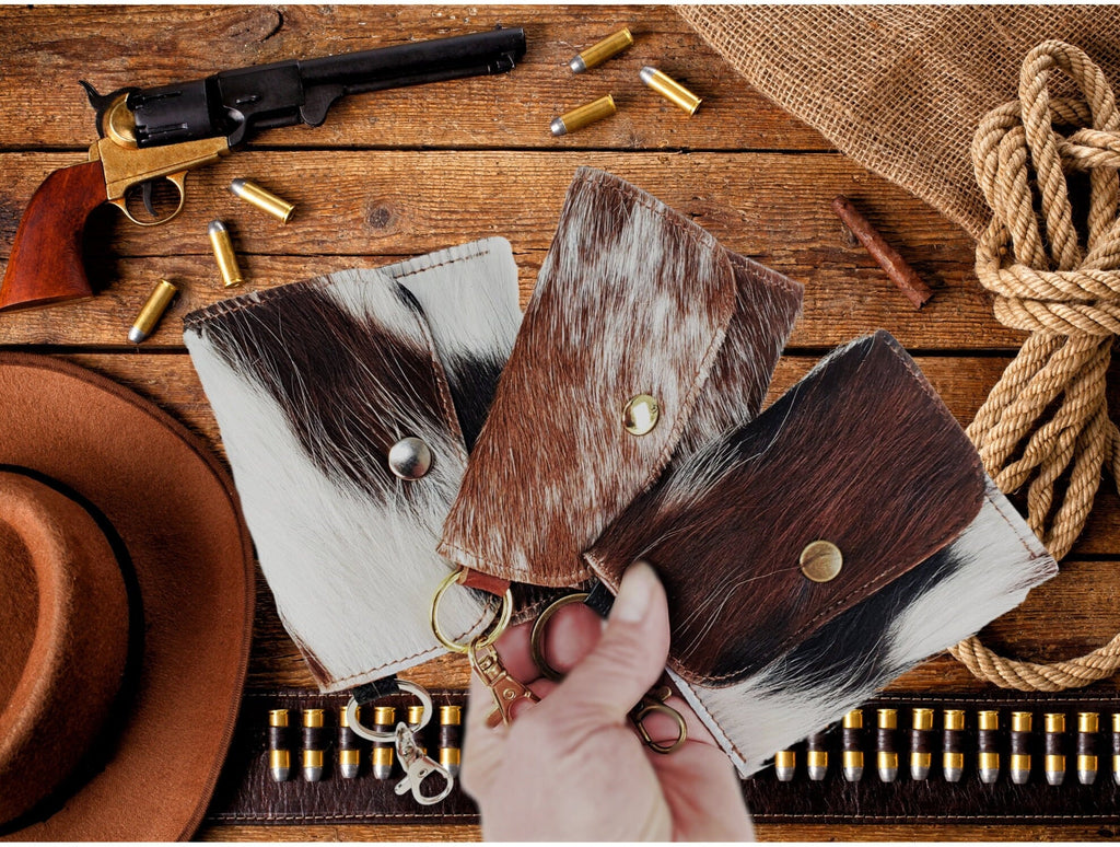 Three hair-on cowhide card wallet keychains in different natural patterns displayed on rustic Western wood